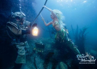 Diver in a restored antique brass diving helmet holding a lantern beside a mermaid on a rocky reef off Santa Catalina Island