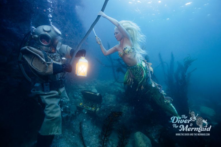 Diver in a restored antique brass diving helmet holding a lantern beside a mermaid on a rocky reef off Santa Catalina Island