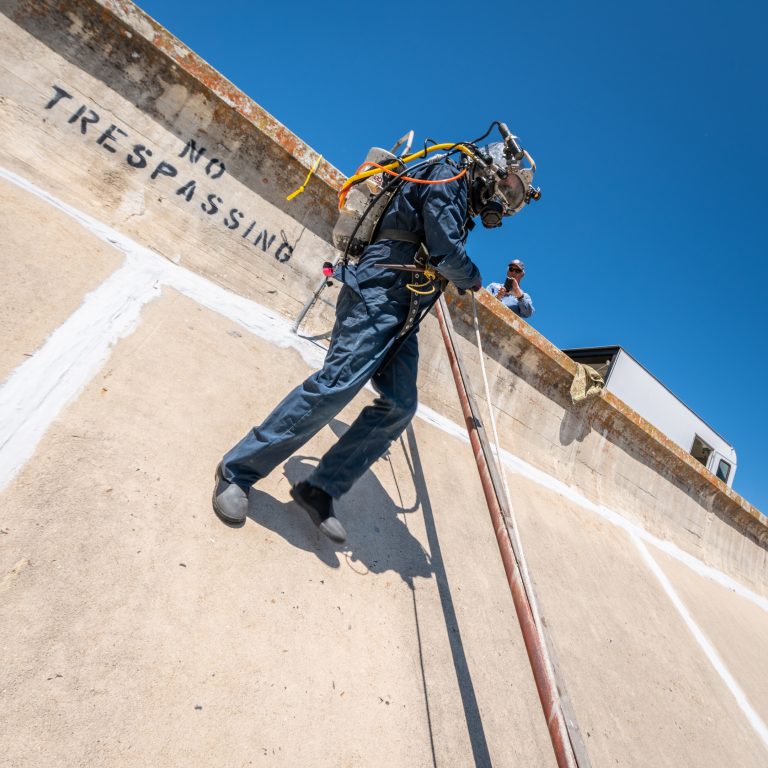 Commercial diver in surface-supplied dive gear climbing down the concrete face of a dam